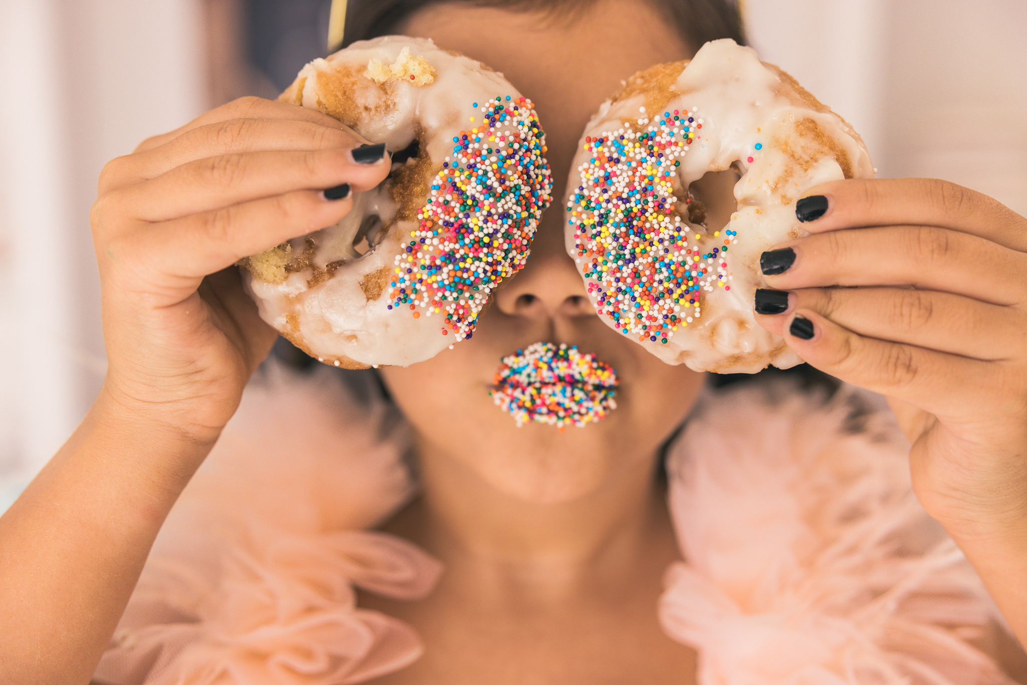 close up of girl in pretty pink dress with sprinkles on her lips and holding two sprinkled donuts over her eyes like glasses in warrenville, il