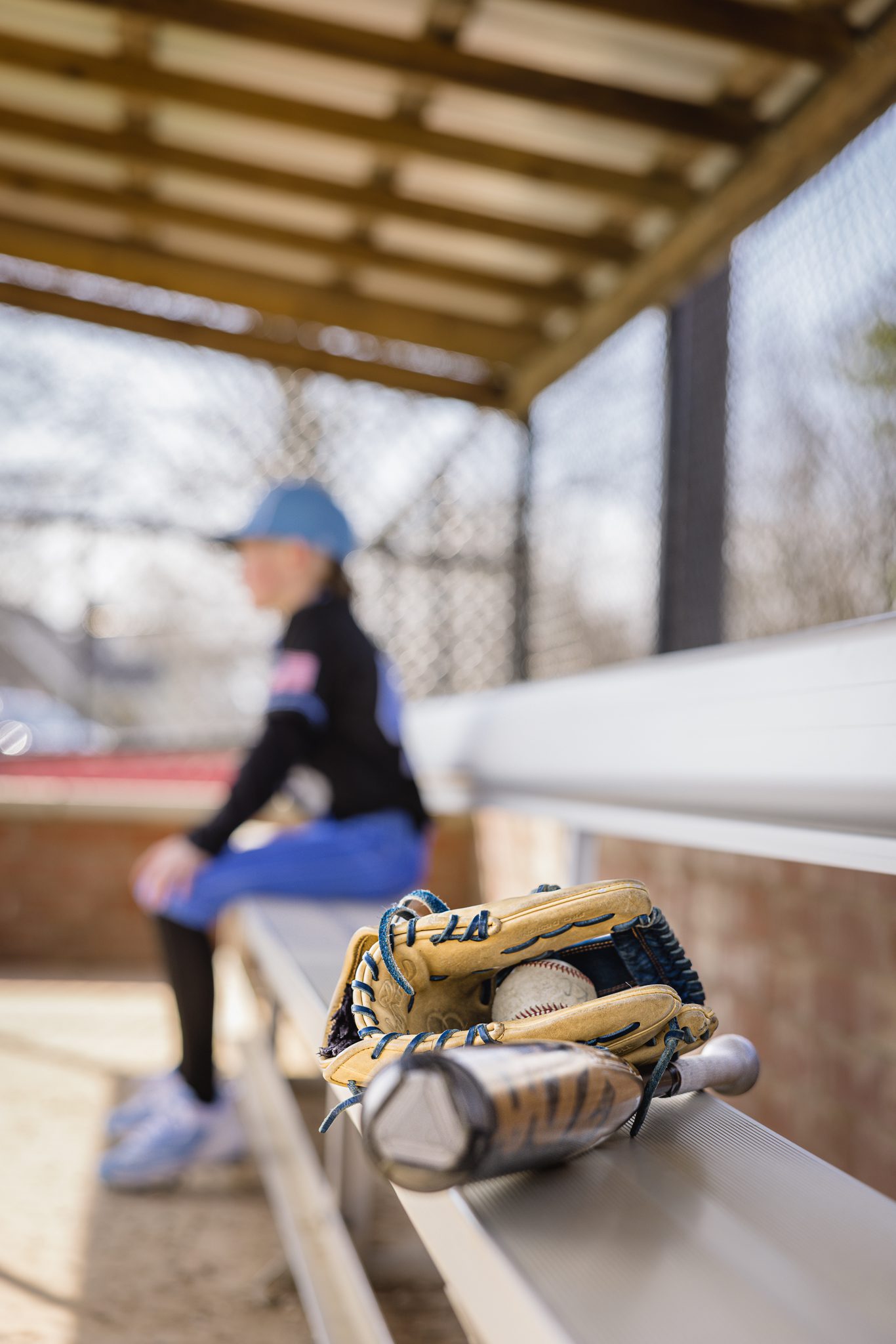 Boy in baseball uniform sitting at end of the bench out of focus while bat, ball and glove close to camera and in focus at Channahon Pony Baseball Fields in Channahon, IL