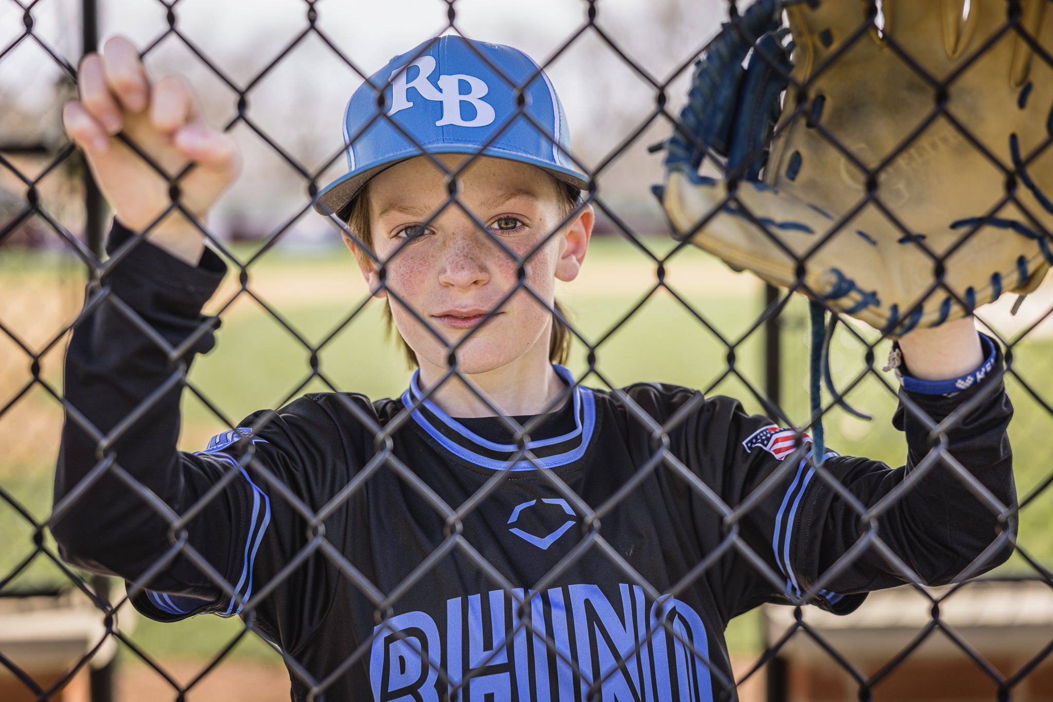 Boy in baseball uniform looking straight into the camera while holding his glove up to his face and standing against a chain length fence at Channahon Pony Baseball Fields in Channahon, IL