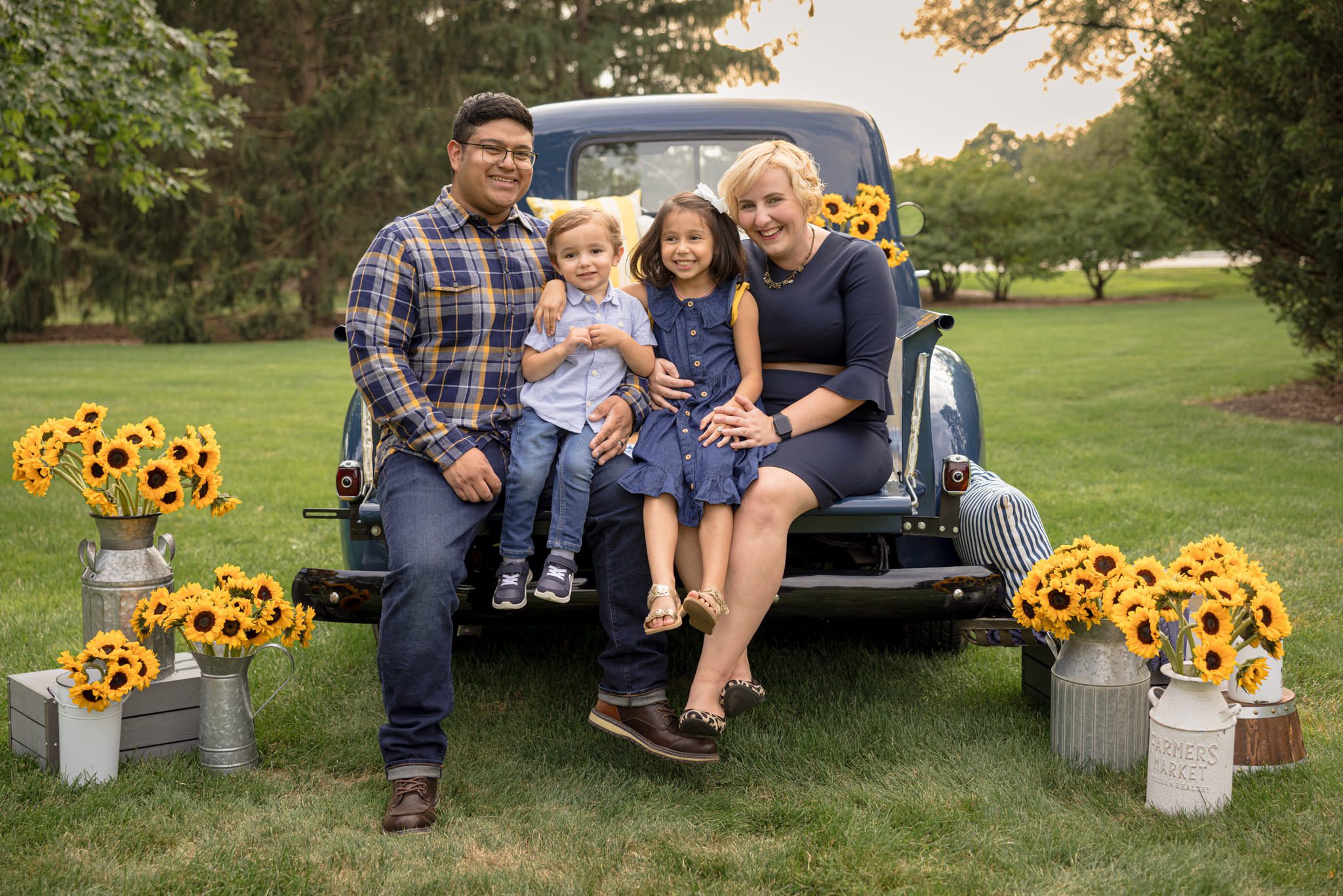 Family with two young children sitting on the back of an antique blue truck that is filled with sunflowers. Photo taken during a family photography session at Danada Equestrian Farm in Wheaton, IL