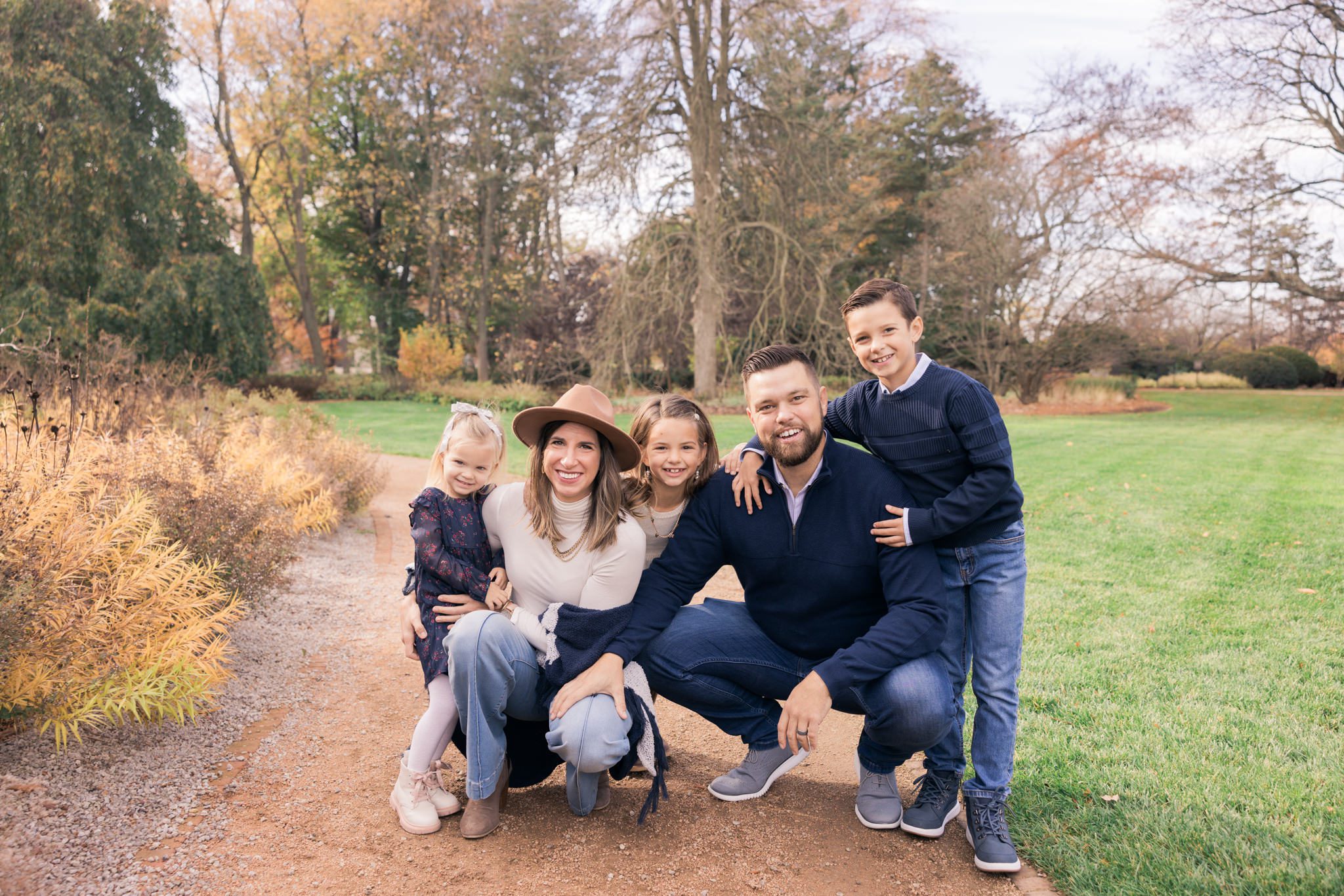 Family with three younger children squatting down on a path and smiling at the camera. Taken during a family photography session at Cantigny in Wheaton, IL