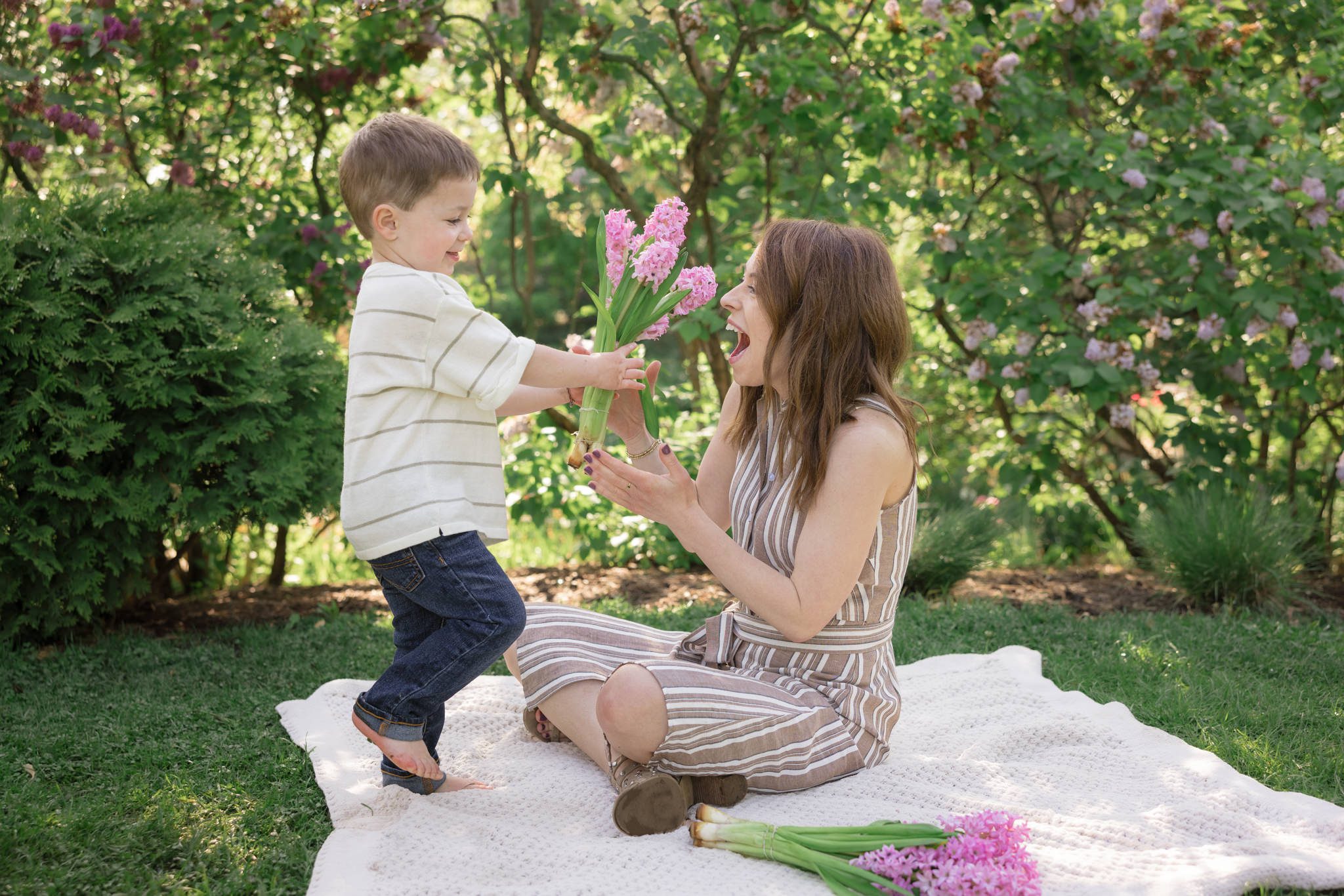 Mom and young son. Mom sitting on blanket while son runs to give mom a bouquet of pink flowers. Taken during a family photography session at Lilacia Park in Lombard, IL