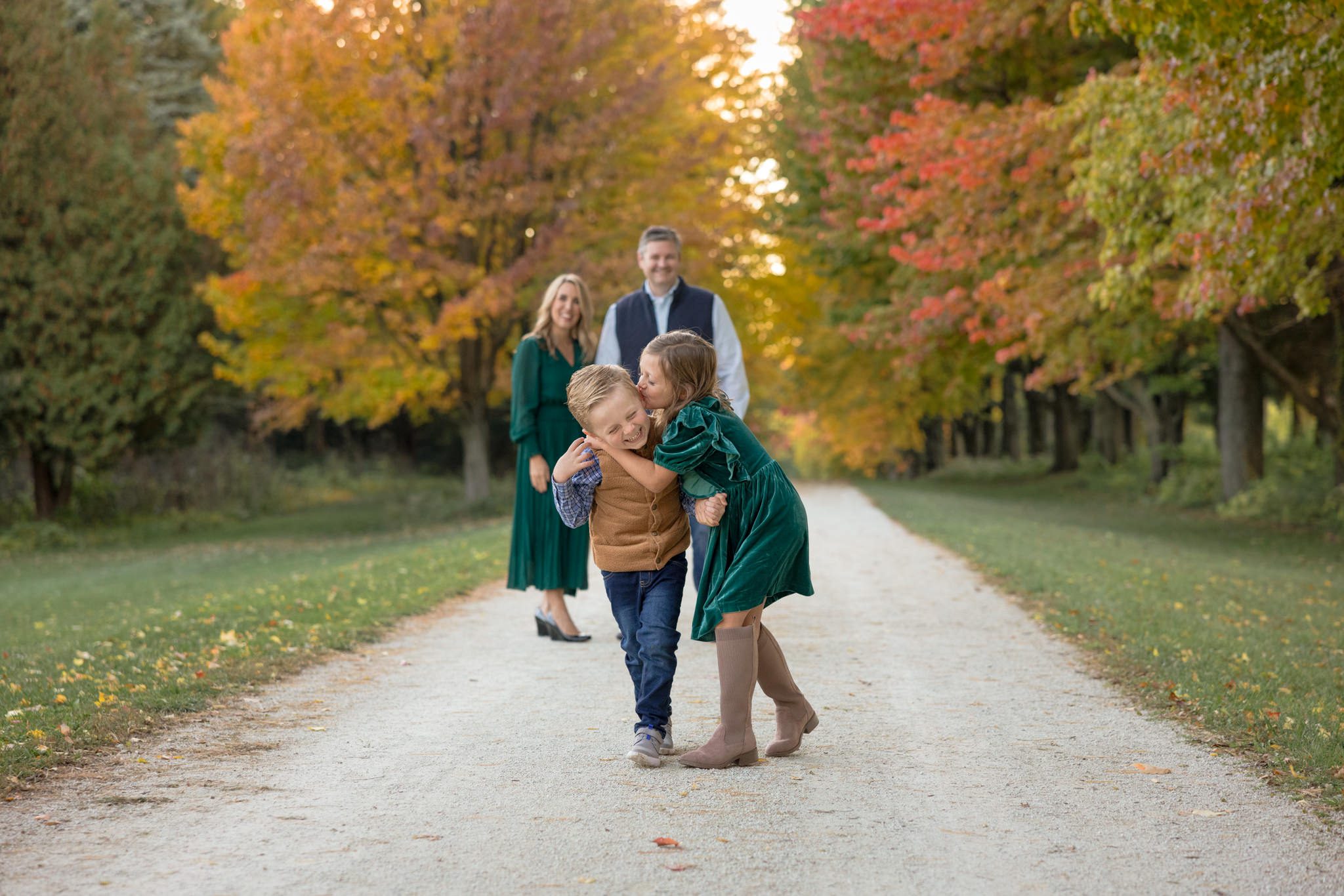 Brother and sister walking down a tree lined path while sister kissing her brother and mom and dad looking on. Taken during a family photography session at St James in Warrenville, IL
