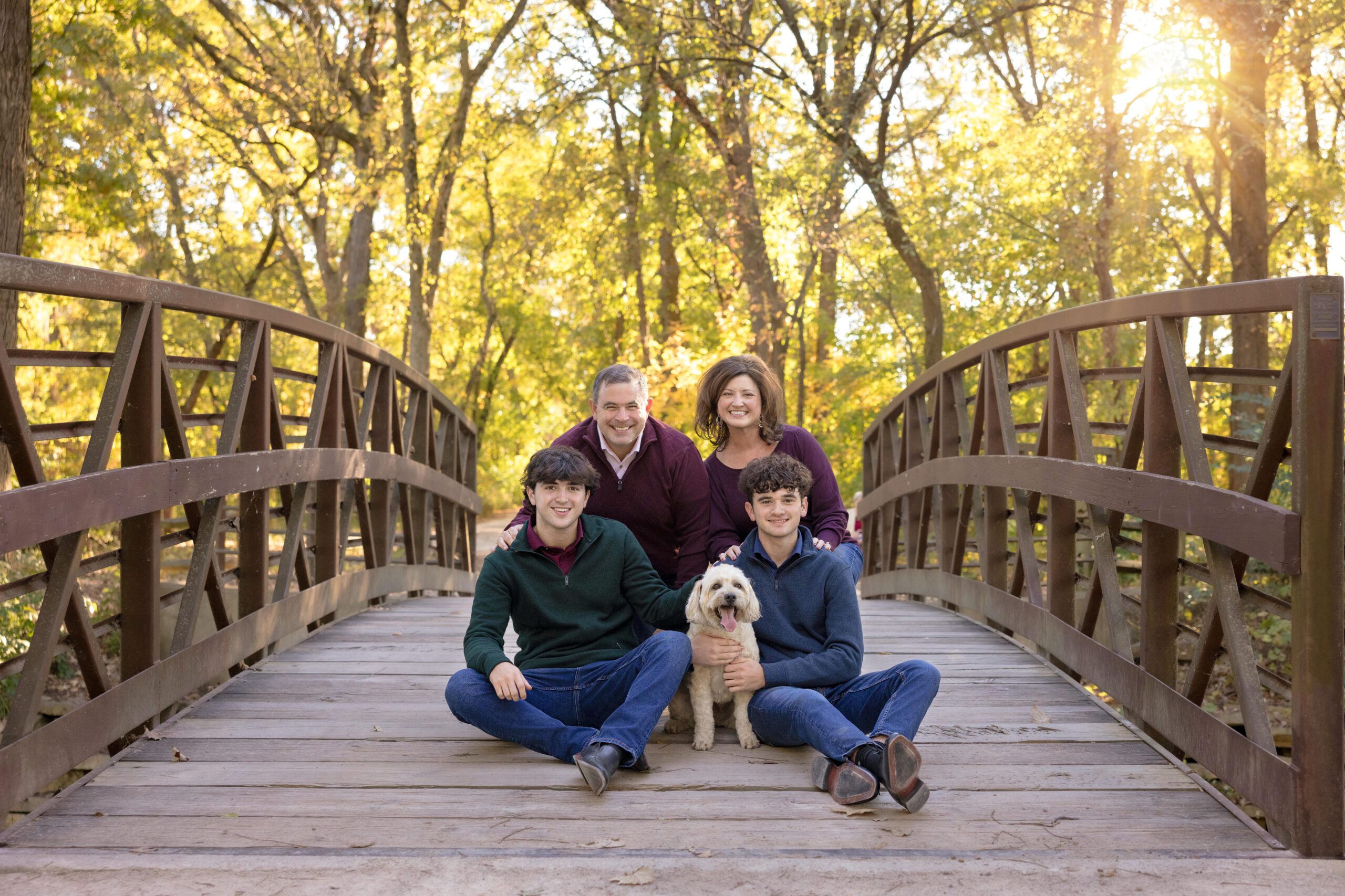 Mom and dad with two older sons and their dog squatting down on a bridge and smiling at the camera. Taken during a family photography session at Herrick Lake in Wheaton, IL