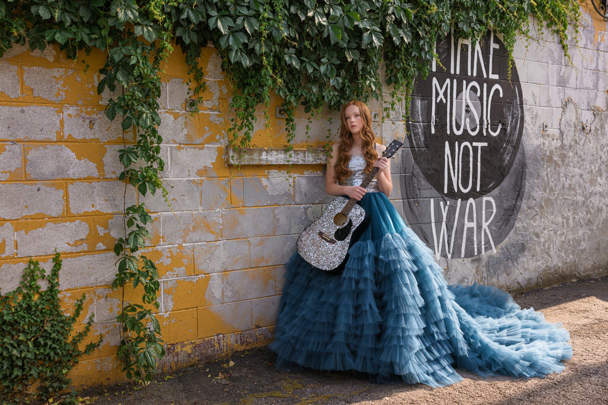 Teenage girl in silver and blue couture dress leaning up a wall with mural and holding a silver guitar. Graduating senior session.