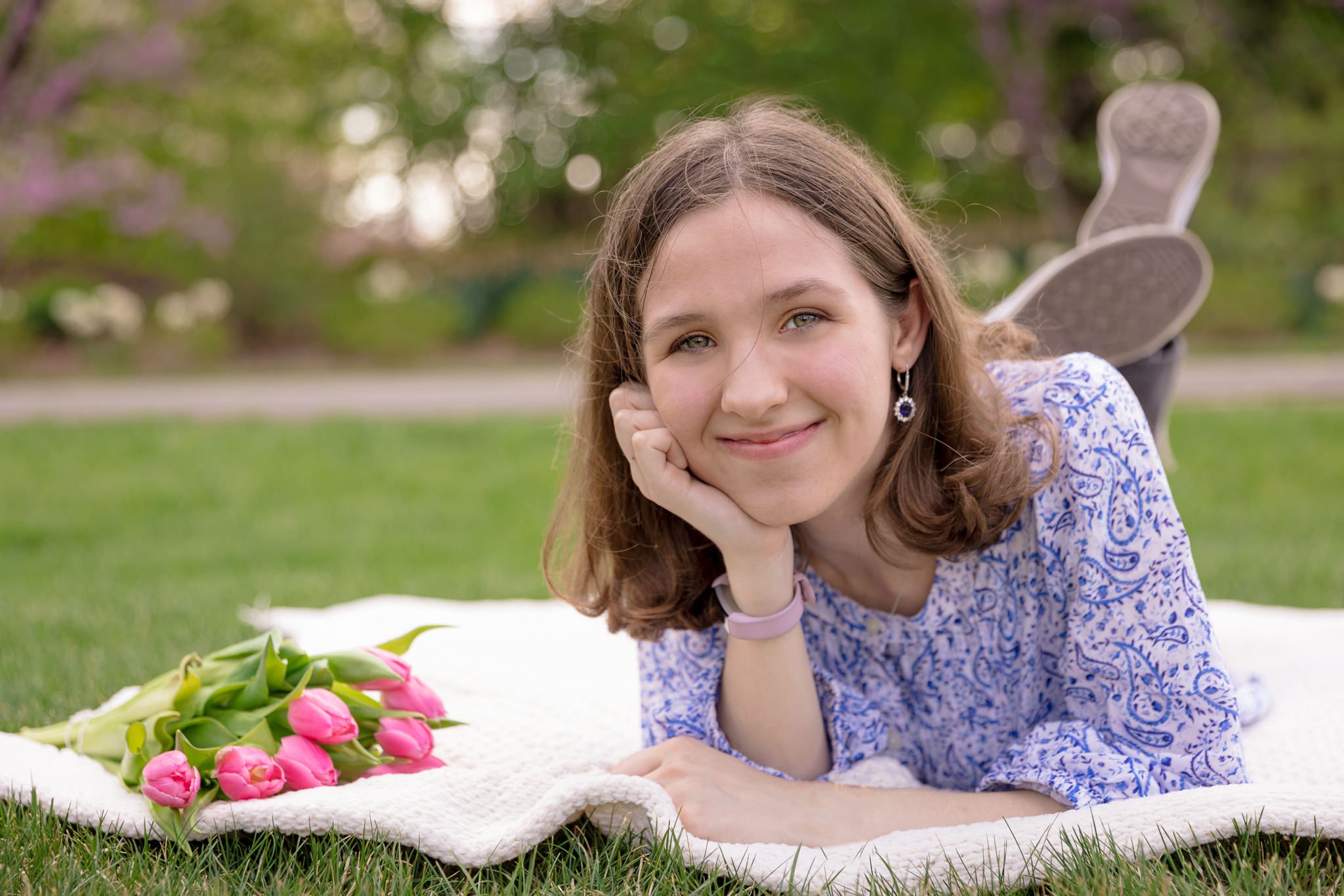 Teenage female lying on a blanket looking and smiling on the camera while a bouquet of pink tulips lying next to her. Photo taken during graduating senior session at Cantigny Park in Wheaton, IL