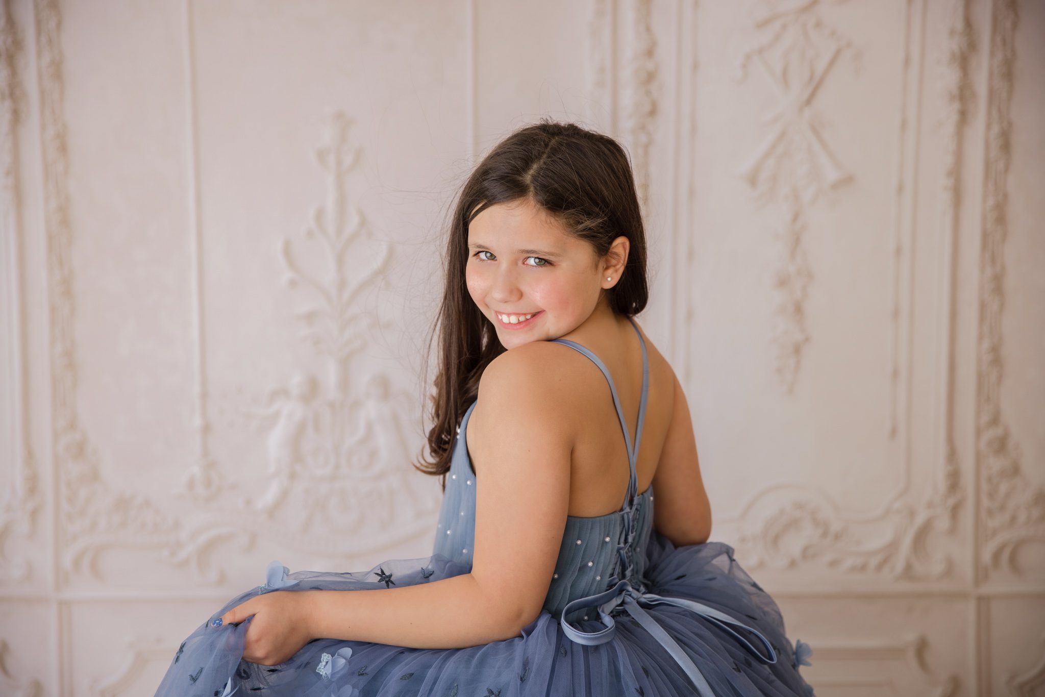 Young girl in a long blue couture dress looking over her shoulder and smiling at the camera. Taken during a studio photography session in Warrenville, IL