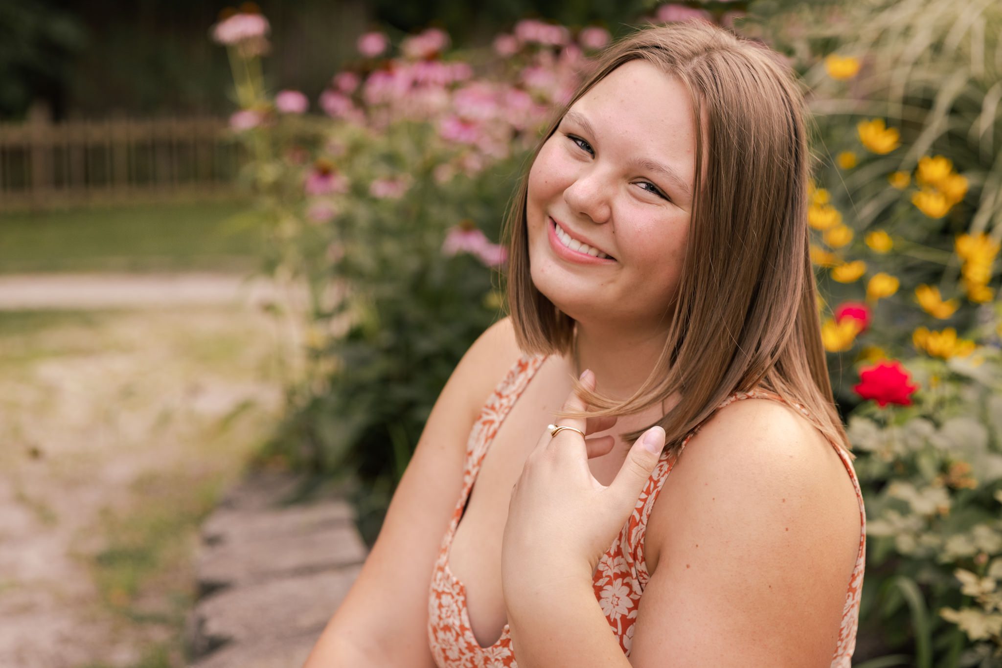 Teenage girl in orange floral dress sitting in front of a bed of summer flowers. Photo taken at Fabyan Park in Geneva, IL